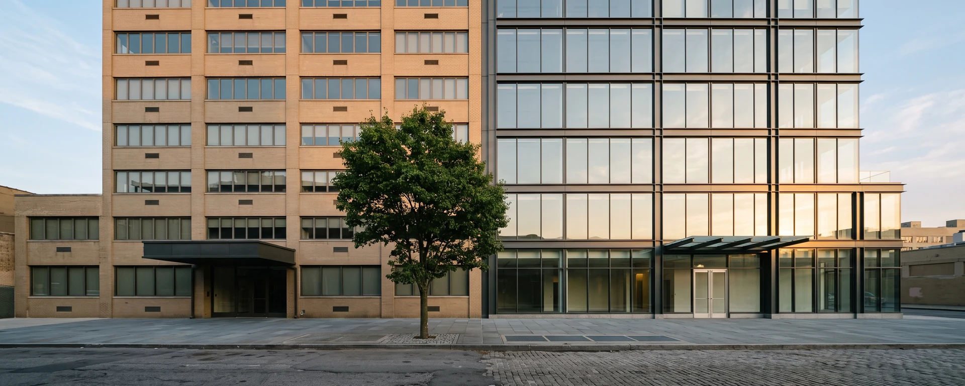 An older brick office building beside a newer glass-and-steel building on a New York City side street in late afternoon light.