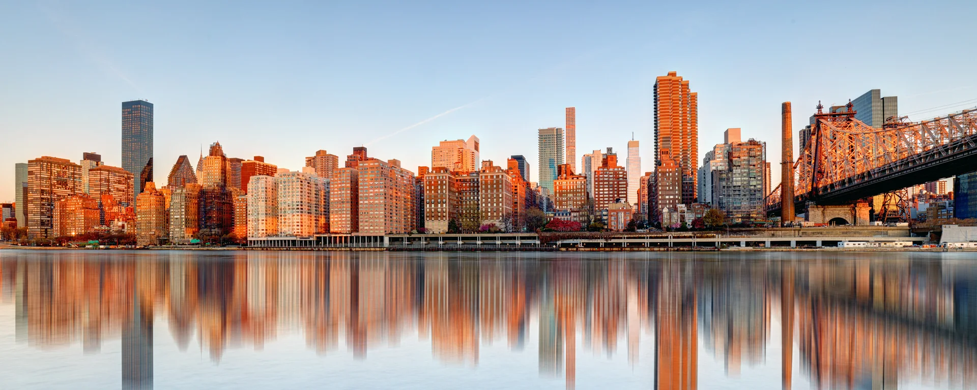 The Queensboro Bridge and the East River skyline at sunrise, mirrored in still water, seen from Queens.